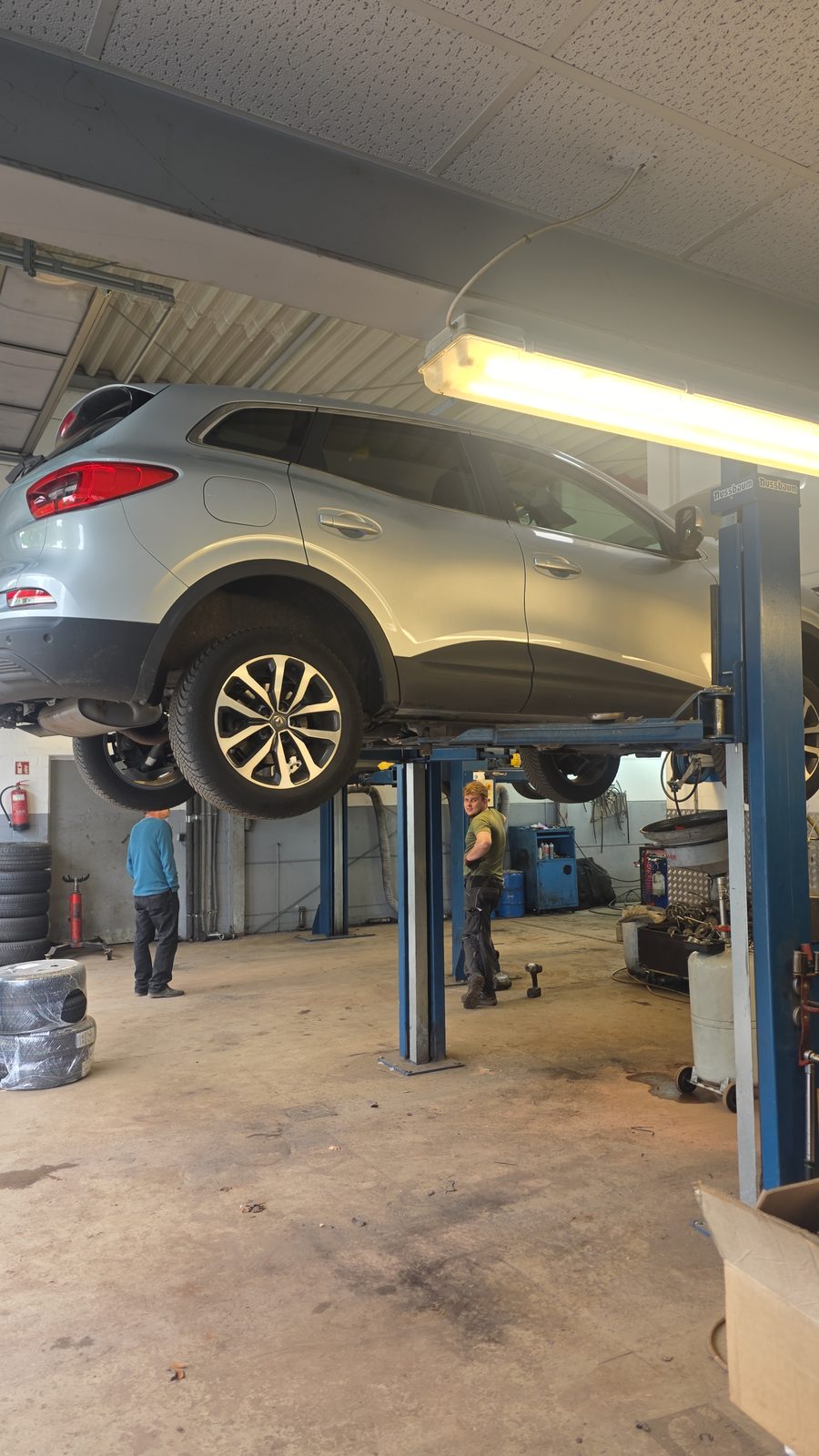 Silberner SUV auf der Hebebühne einer Kfz-Werkstatt, während zwei Mechaniker an der Reparatur arbeiten