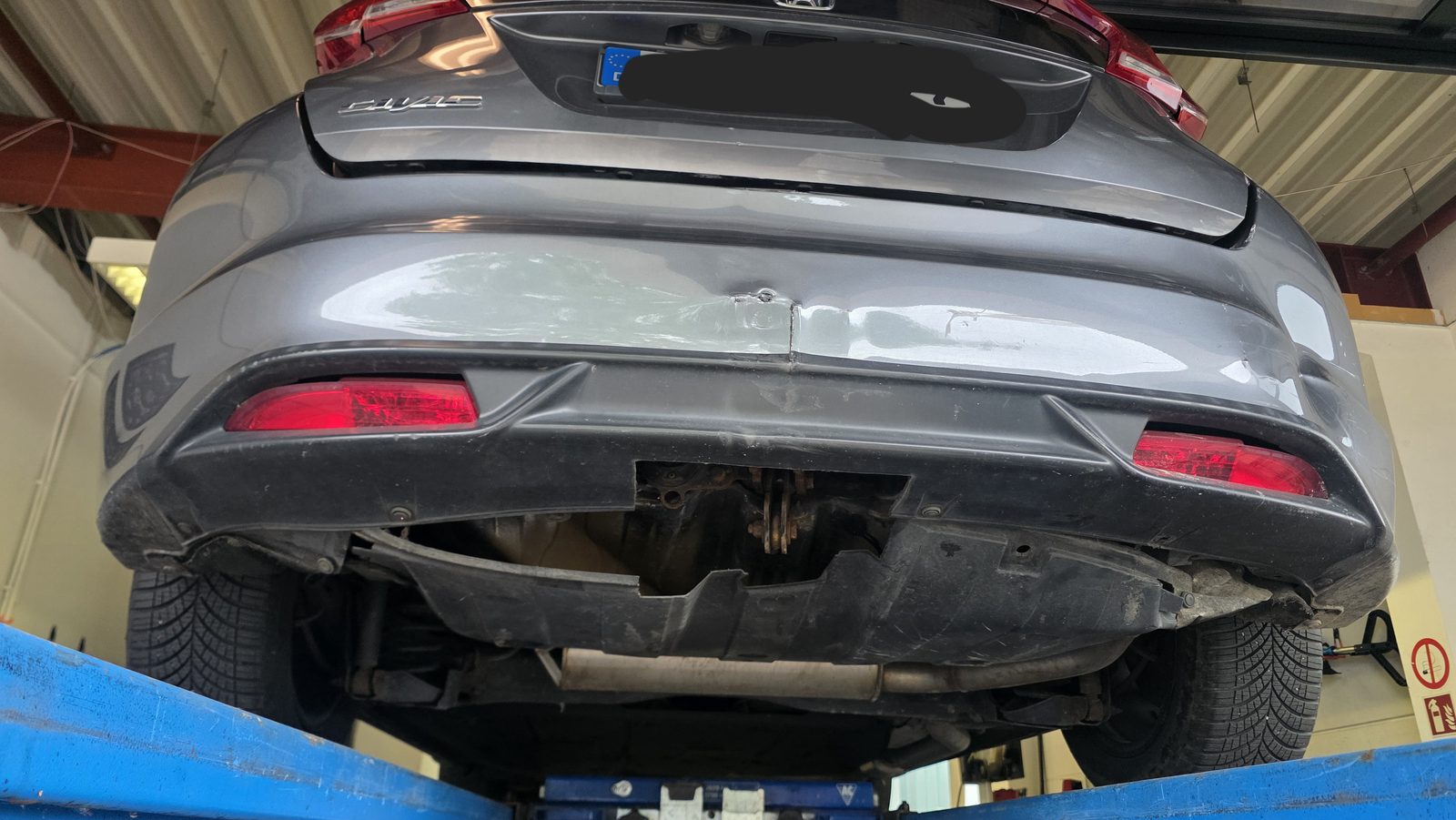 Graue Honda Civic auf der Werkstattbühne mit Heckschaden am Stoßfänger von unten begutachtet